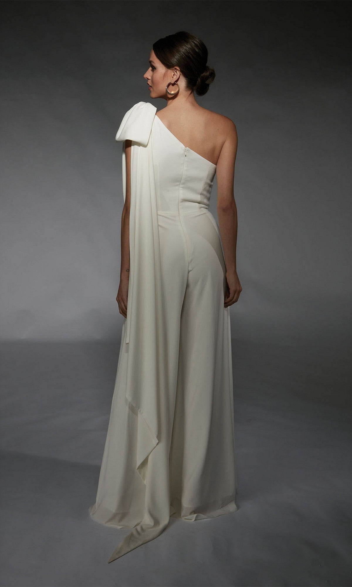 A woman stands facing away against a gray background in the Alyce 70065—an elegant white, one-shoulder long formal jumpsuit with draped detail, offering a chic alternative to a cocktail dress.